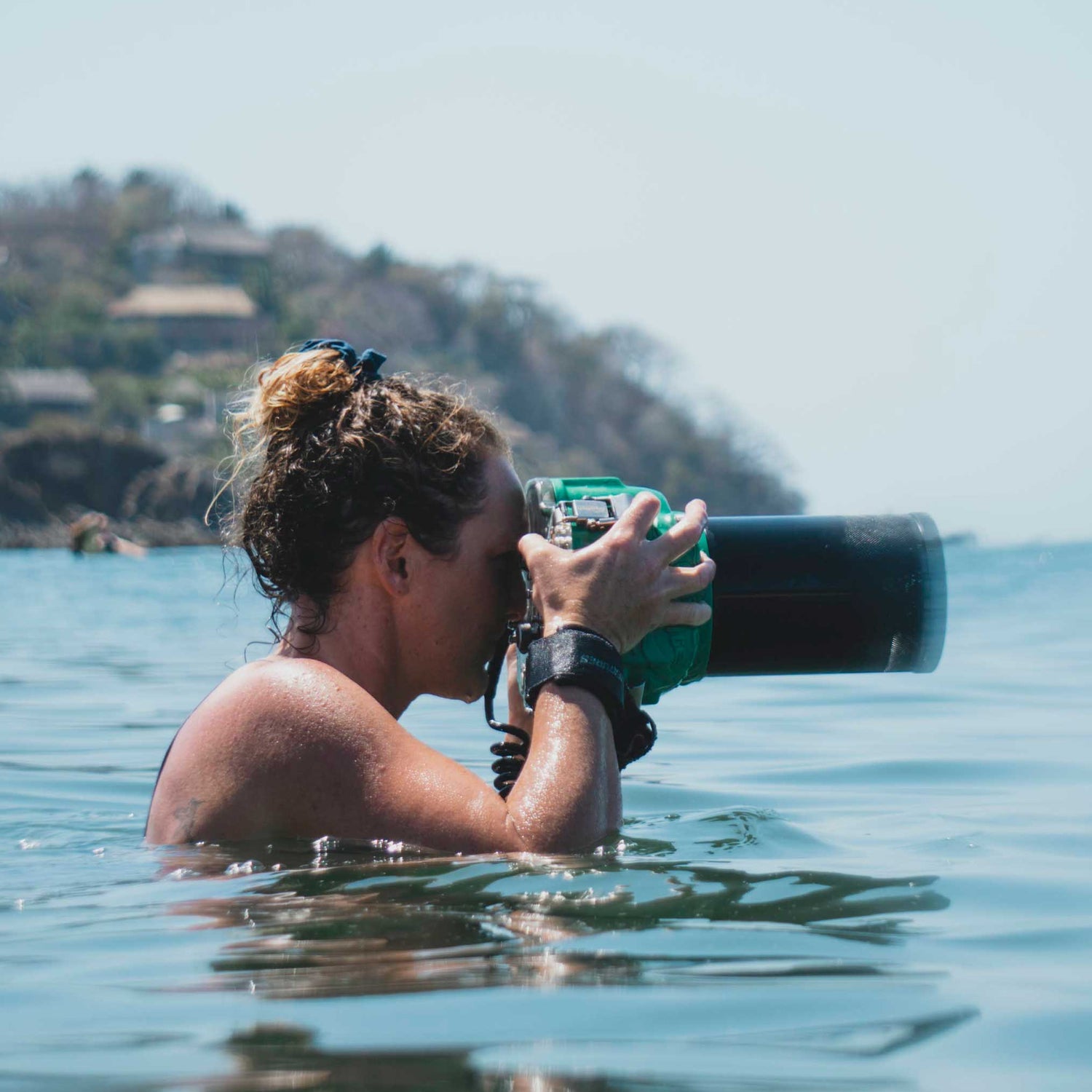 Person holding a camera underwater in clear blue water with a scenic background