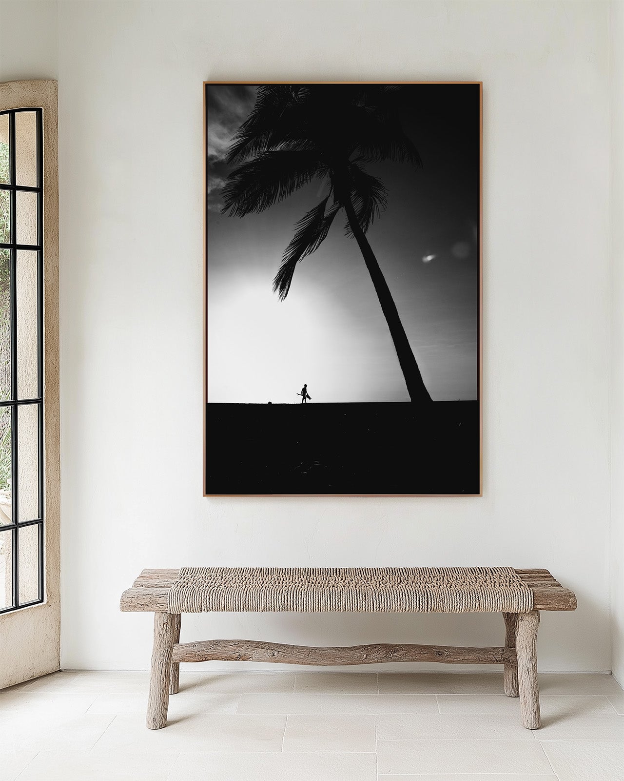 Framed black and white artwork of a palm tree and person on a beach, displayed in a minimalistic room with a wooden bench.